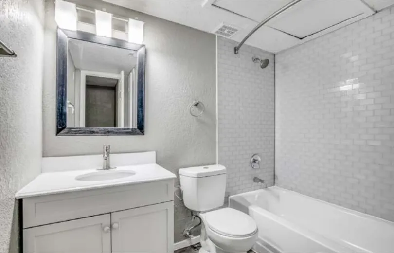Apartment bathroom with a white vanity, a framed mirror with a light fixture above it, a toilet, and a bathtub with a tiled surround and curved shower rod.