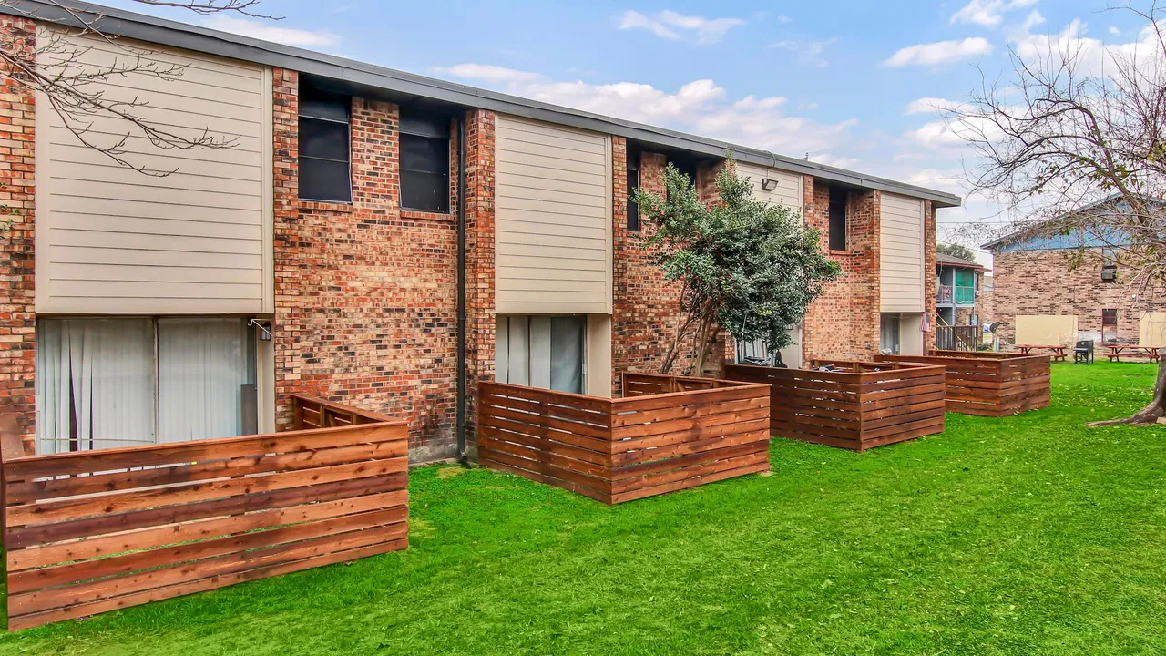 A row of two-story brick and siding apartment buildings with private wood-fenced patios facing a green lawn.