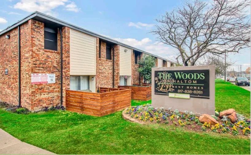 Two-story brick and siding apartment buildings with wood-fenced patios, and the Woods of Haltom community sign standing in a flower bed on a green lawn.