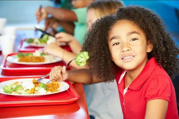 A smiling girl in a red shirt sitting in front of a school lunch tray with food.