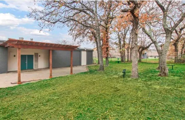 A grassy yard with large trees and a wooden covered pergola in front of a low building with teal doors.